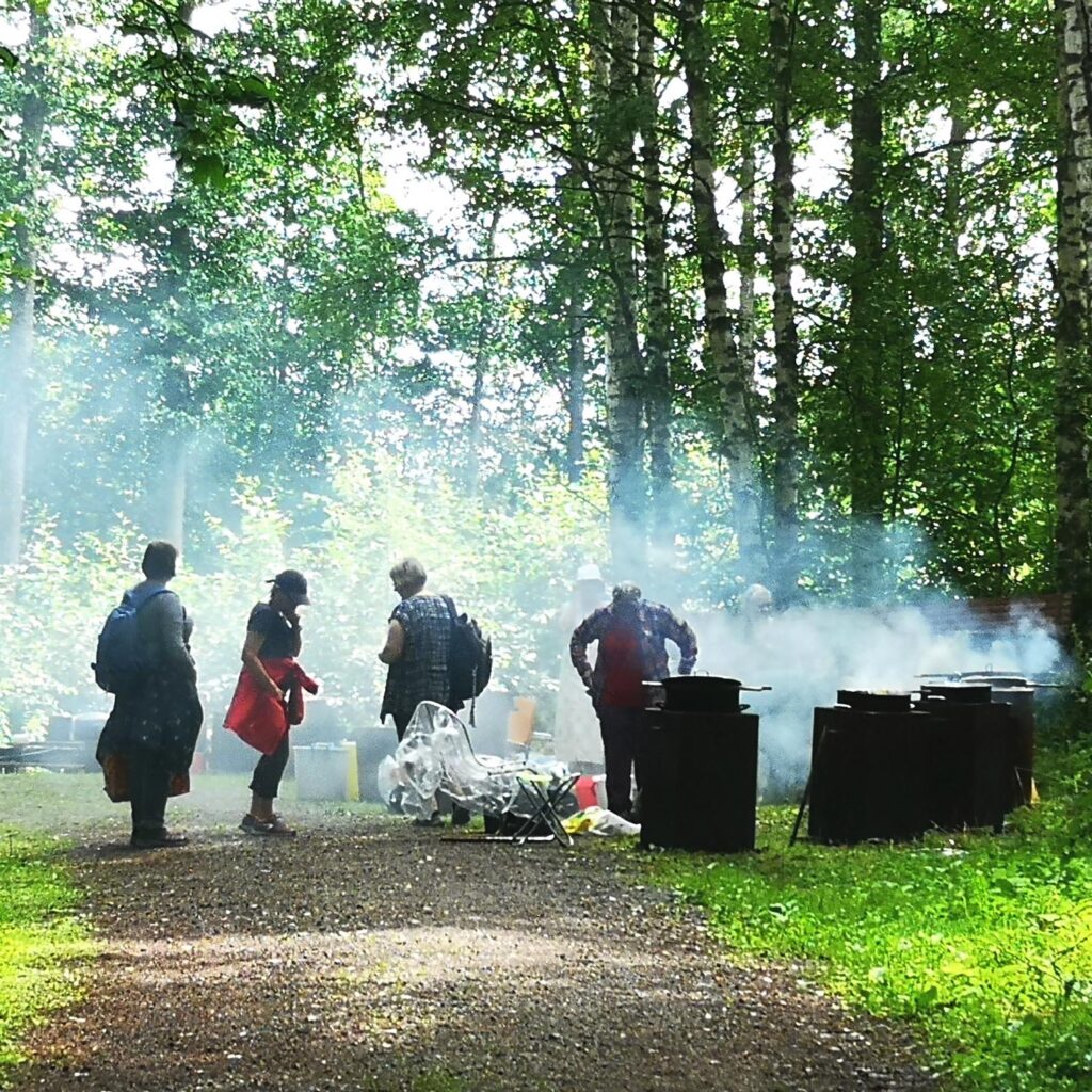 Yhdistysnaisia höyryävien kattiloiden keskellä metsässä.